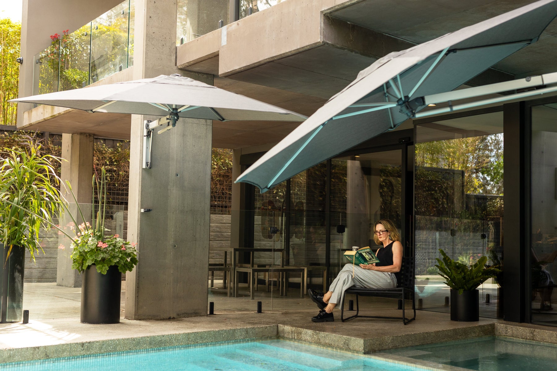 Woman sitting down and reading by the pool where she has two wall mounted cantilever umbrellas by Shadowspec providing shade