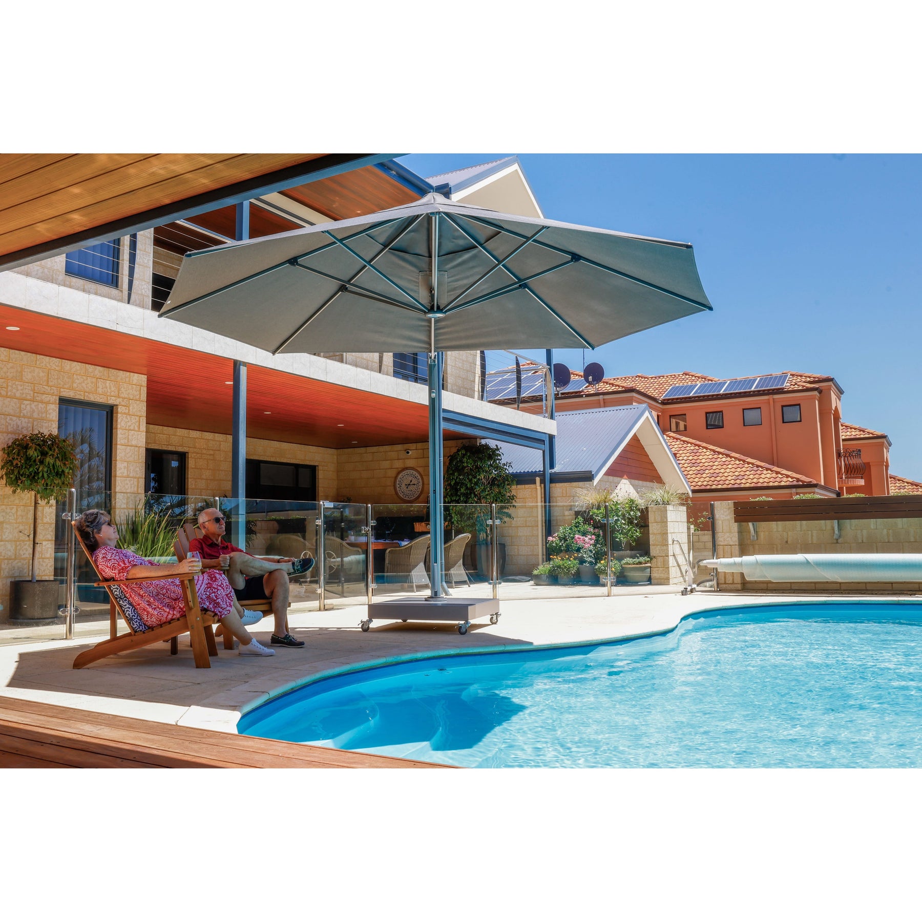 A large grey cantilever patio umbrella shading a couple relaxing on wooden lounge chairs next to a bright blue swimming pool with a house in the background.