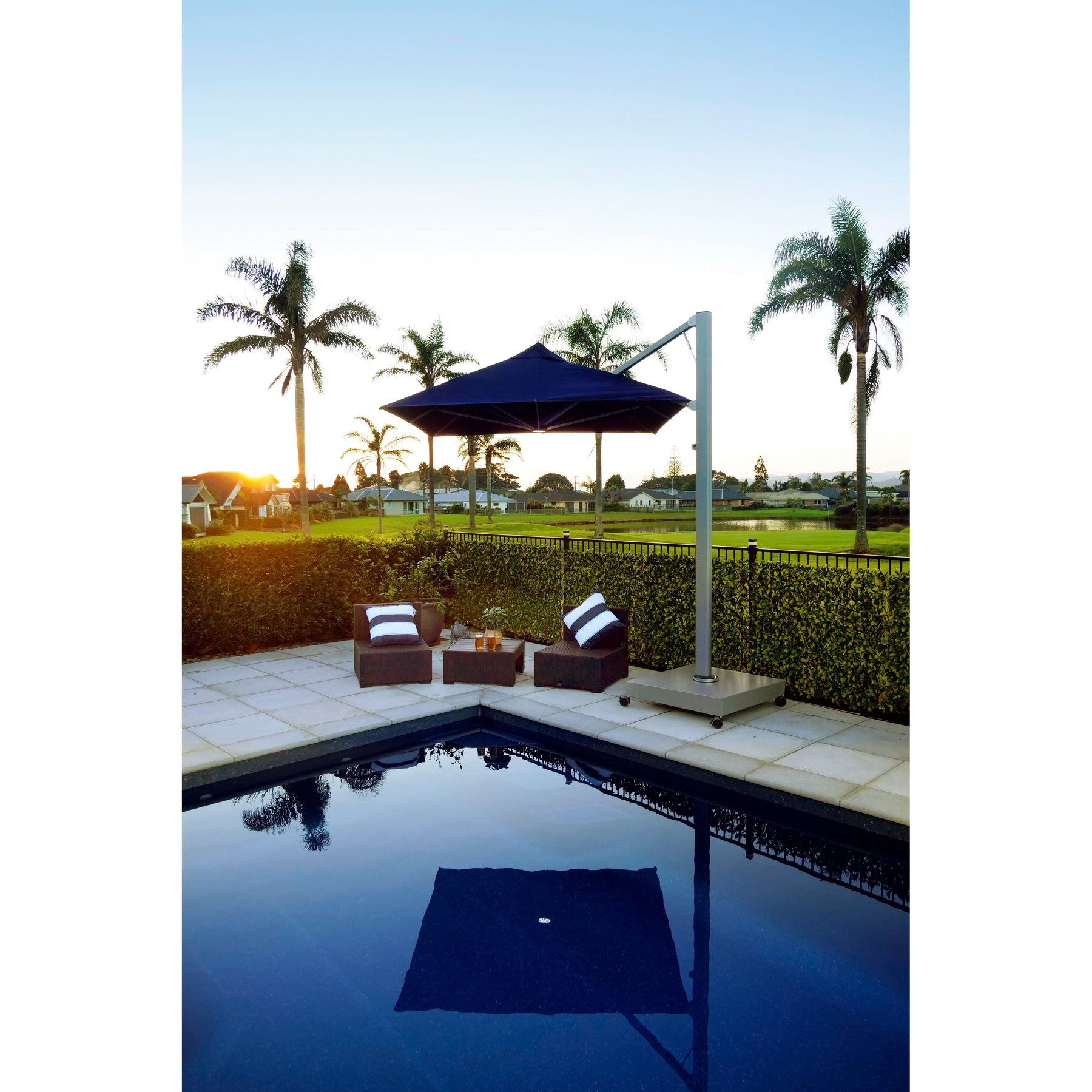 A Shadowspec Serenity dark blue square cantilever umbrella shading two wicker lounge chairs on a patio beside a swimming pool.