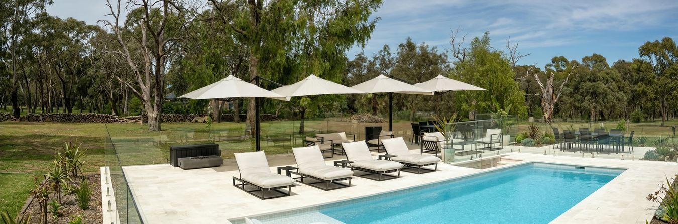 A wide panoramic view of a luxury pool area featuring four large white commercial cantilever umbrellas providing shade over premium sun loungers.