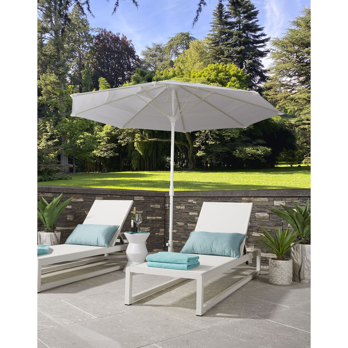 White Helux patio umbrella over two lounge chairs with blue cushions on a stone patio with greenery in the background.