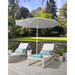 White Helux patio umbrella over two lounge chairs with blue cushions on a stone patio with greenery in the background.
