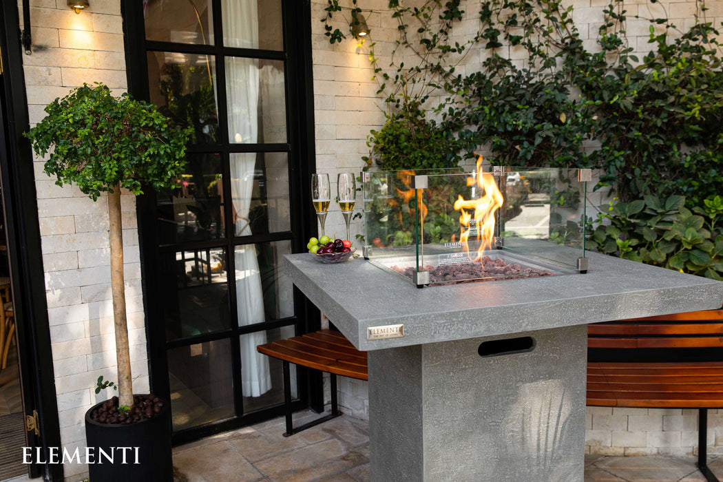 Outdoor patio with Elementi Montreal fire pit table, plants, and a modern building entrance.