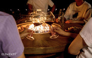 People around an Elementi Lafite fire pit table with glasses of wine, enjoying a warm evening together.