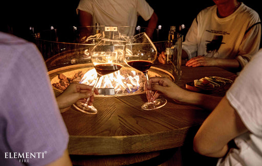 People around an Elementi Lafite fire pit table with glasses of wine, enjoying a warm evening together.
