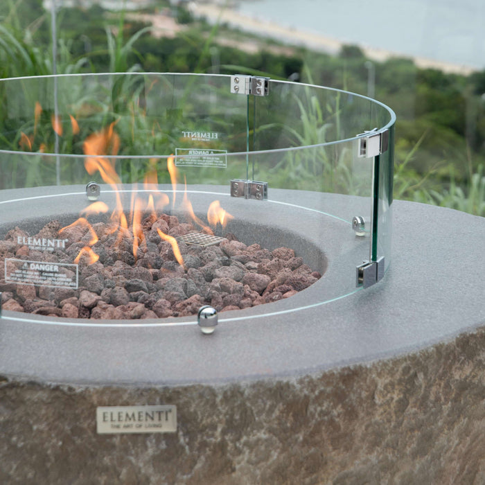 Elementi Boulder Fire pit with glass enclosure and visible flames, set against a scenic background.