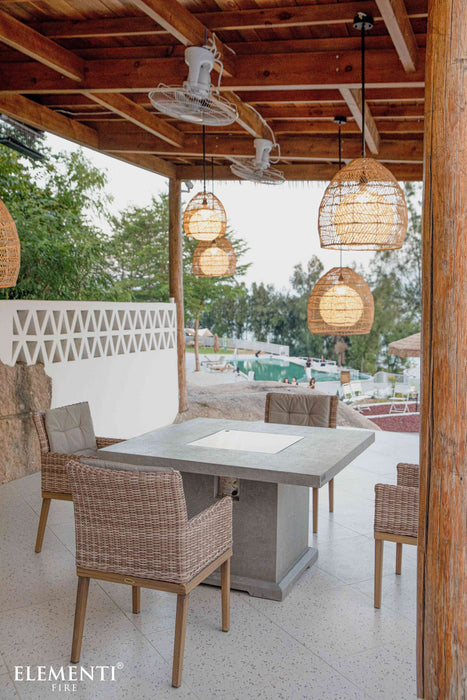 Outdoor dining area with wicker chairs and an Elementi fire table under a wooden pergola, featuring the Elementi brand.