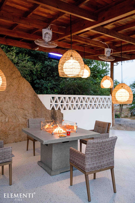 Outdoor patio with an Elementi Birmingham dining and fire table, chairs, and hanging lights in a garden setting.