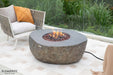 Round stone fire pit table with flames on a patio, surrounded by outdoor furniture.
