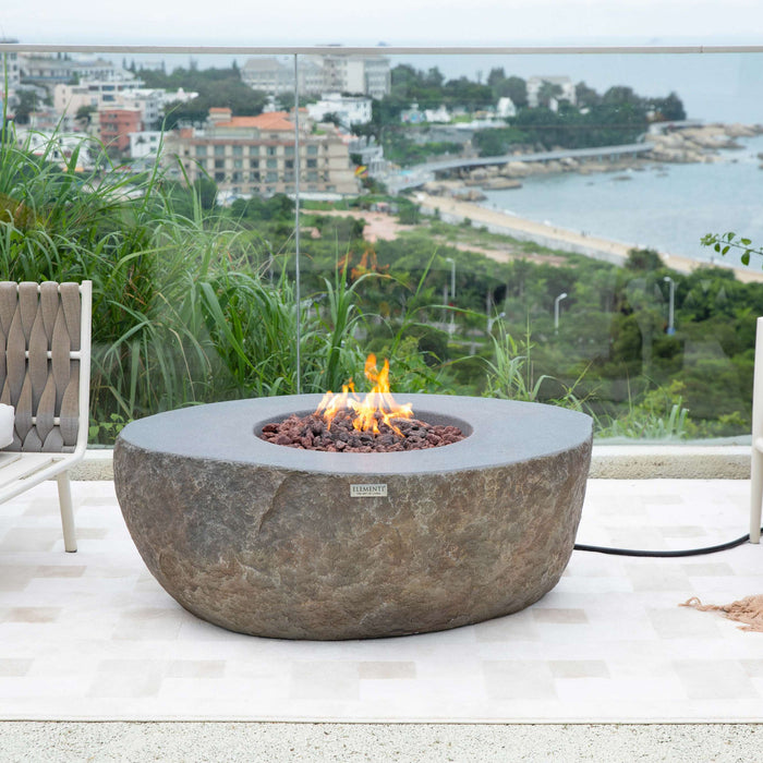 Fire pit table with flames on a patio overlooking a scenic view with chairs and a blanket.