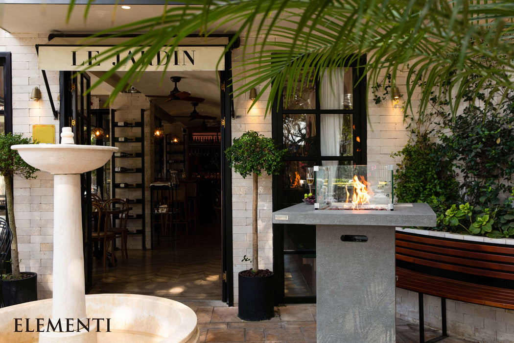 Outdoor setting with a fountain, bench, Elementi Montreal fire table, and building entrance.