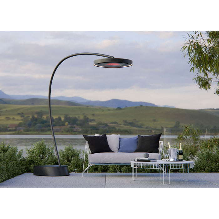 Outdoor setting with a modern black Bromic Eclipse Portable heater doubling as a floor lamp, white sofa, and small table by a scenic landscape.