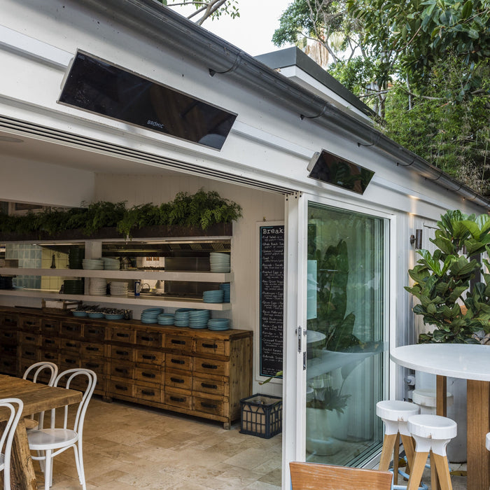 Modern kitchen with open sliding door, wooden cabinets, two Bromic Platinum Heaters mounted above the sliding door, and white stools.