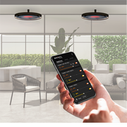 A person holding a smartphone displaying the Bromic Heating App interface, using it to adjust the settings of two circular, ceiling-mounted Eclipse Electric heaters in a modern outdoor patio setting