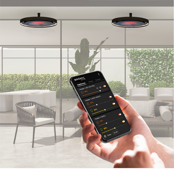 A person holding a smartphone displaying the Bromic Heating App interface, using it to adjust the settings of two circular, ceiling-mounted Eclipse Electric heaters in a modern outdoor patio setting