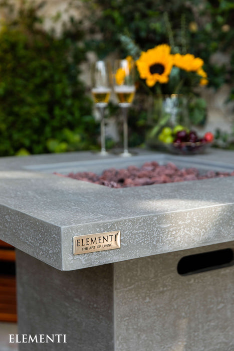 Concrete fire pit table with 'Elementi' branding, glasses of wine, and flowers in the background.