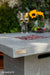Concrete fire pit table with 'Elementi' branding, glasses of wine, and flowers in the background.