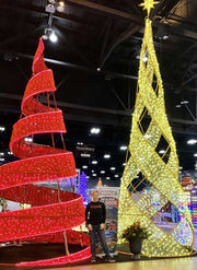 Person standing between a red spiral and yellow geometric Christmas tree decoration indoors.