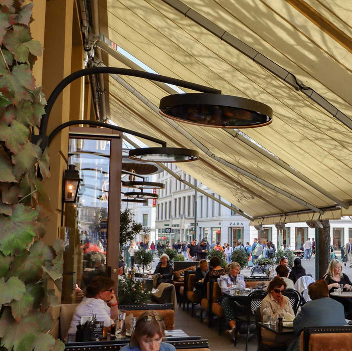 People dining outdoors under a large canopy with street view in the background and the focus placed on the Bromic Eclipse Heaters that are wall mounted
