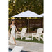 Woman in a white dress walking near two white Adirondack chairs and a Helux umbrella on a patio.