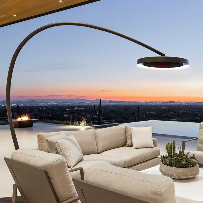 Outdoor patio with beige sofa, armchair, and a Bromic Eclipse Portable heater floor lamp overlooking a cityscape at sunset.