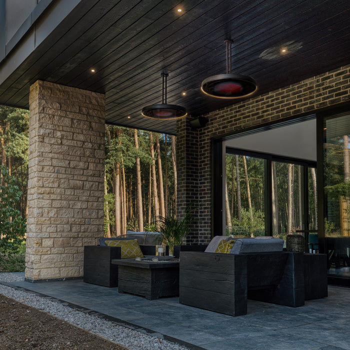 Modern outdoor patio with dark furniture, Bromic Eclipse Heaters hanging from the ceiling, and stone wall, surrounded by trees.