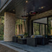 Modern outdoor patio with dark furniture, Bromic Eclipse Heaters hanging from the ceiling, and stone wall, surrounded by trees.