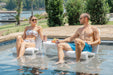 Two people sitting in a pool with drinks, surrounded by greenery and water.
