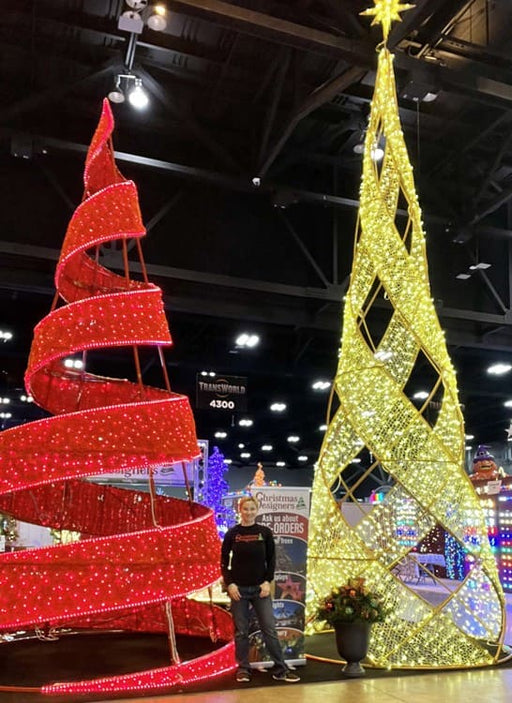 Person standing between a red spiral and yellow geometric Christmas tree decoration indoors.