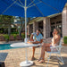Two people sitting under a blue patio umbrella by a poolside.