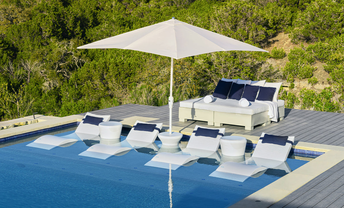 Outdoor lounge area with white chairs, blue cushions, and a large umbrella by a pool with greenery in the background.