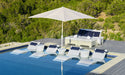 Outdoor lounge area with white chairs, blue cushions, and a large umbrella by a pool with greenery in the background.