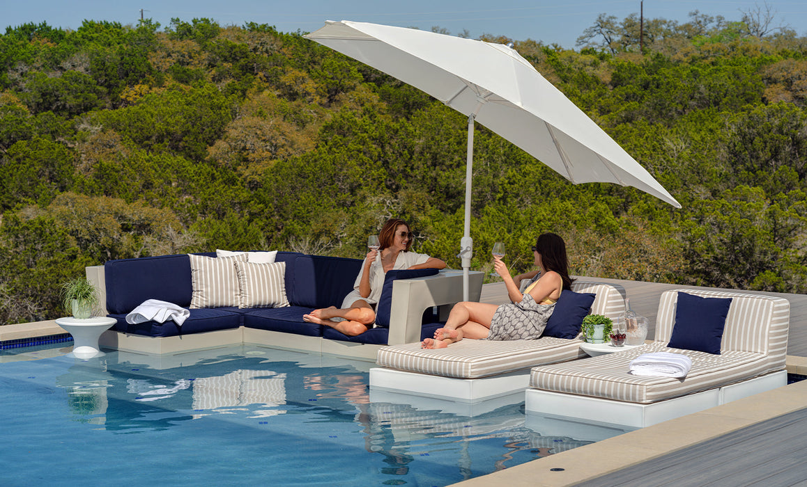 Two women relaxing on a patio with a pool, outdoor furniture, and a tilted ledge lounger umbrella.