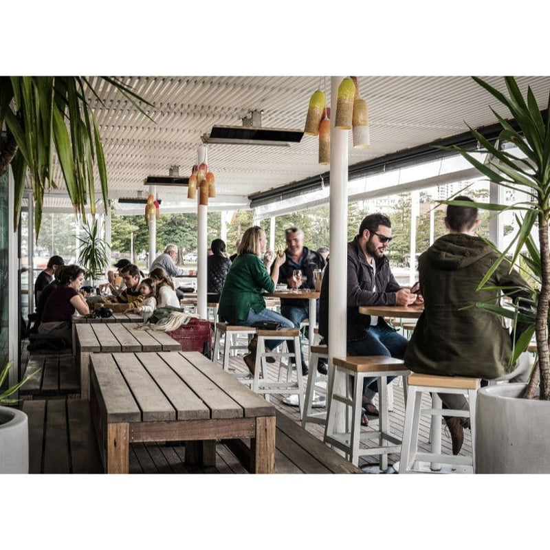 People sitting at tables in an outdoor restaurant setting that has Bromic Heaters mounted to the ceiling with plants and wooden furniture.