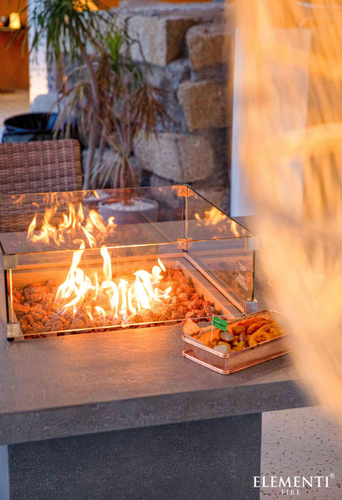 Outdoor fire pit with flames and a blurred person in the foreground, featuring the Elementi Fire brand.