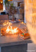 Outdoor fire pit with flames and a blurred person in the foreground, featuring the Elementi Fire brand.
