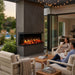 People sitting around an outdoor 3 sided Orion fireplace with a pool and garden in the background