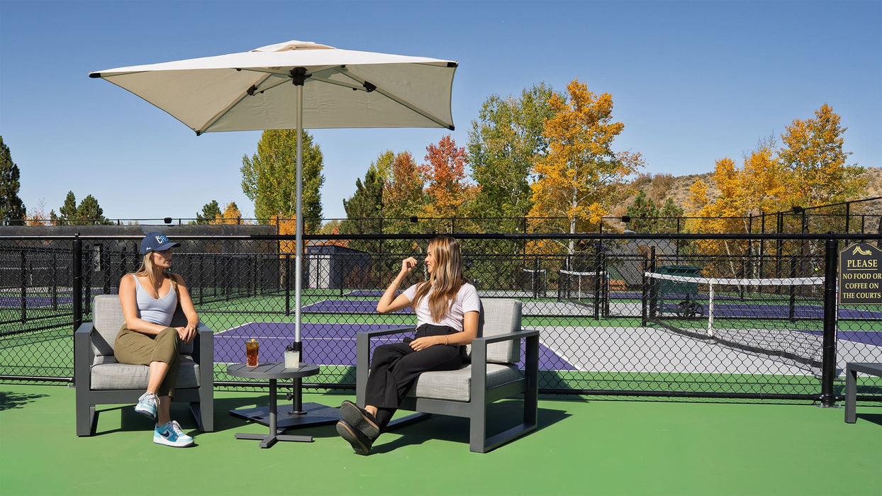 Two women sitting under a beige umbrella on a tennis court with autumn trees in the background.