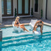 A man dunking the ripple side table by TenJam into the pool water to fill it up while a woman sits behind him on a TenJam Mibster chair on the baja shelf of their pool.