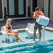 Man pouring water from a TenJam Ripple side table into a pool while a woman sits nearby on a white TenJam Mibster chair