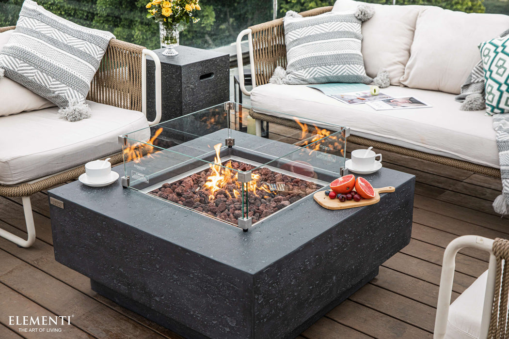 Outdoor patio setting with Elementi Manhattan fire pit table, chairs, and cushions on a wooden deck.