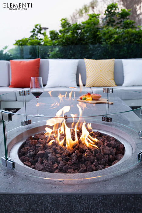Close up of Elementi Metropolis Fire pit table that has a glass wind guard, on an outdoor patio with a couch and colorful cushions.