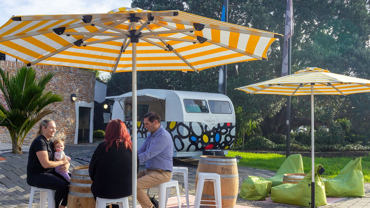 People sitting under yellow and white striped umbrellas with a colorful van in the background.
