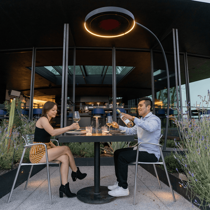 Two people sitting at a table in an outdoor restaurant setting, enjoying drinks while a Bromic Eclipse Portable patio heater hangs above their table