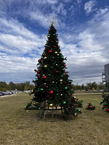 Decorated Christmas tree with colorful ornaments on a grassy area under a blue sky with clouds.