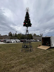 Decorated Christmas tree on a stand in an outdoor setting with cars and buildings in the background.