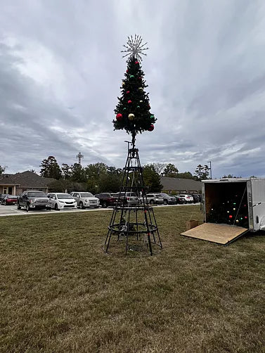 Decorated Christmas tree on a stand in an outdoor setting with cars and buildings in the background.